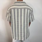 Camisa de manga corta, cuello cubano, color crudo con rayas verticales finas en gris oscuro. Presenta un bolsillo de parche en el pecho y botones marrones.
