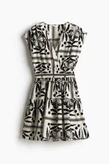 Vestido corto sin mangas de algodón color crudo con estampado de flores, hojas y lunares en negro. Tiene escote cruzado en V, cintura elastizada y falda con vuelo.