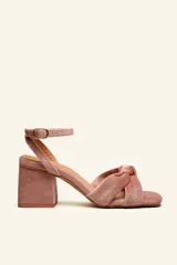Sandalia de tacón color rosado, confeccionada en terciopelo. Presenta una tira ancha anudada en la parte delantera y una tira fina con hebilla dorada alrededor del tobillo. El tacón es cuadrado y de 7 cm de alto.