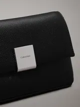 Cartera cruzada negra de piel sintética granulada, con correa de cadena plateada y cierre de solapa con placa metálica con el logo de Calvin Klein grabado.