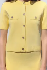 Chaqueta de punto color amarillo pálido, con cuello redondo y manga corta. Cierre frontal con botones dorados y bolsillos de parche en el pecho.