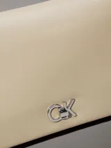 Cartera tipo sobre de piel sintética color beige claro, con solapa frontal y herrajes plateados. Presenta el monograma 'CK' en metal en la parte frontal y una correa de cadena convertible que permite usarla al hombro o cruzada.