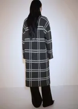 Abrigo largo de punto grueso con estampado de cuadros grises sobre fondo negro. Tiene cuello con solapa, cierre frontal con botones y dos bolsillos de parche.