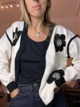 Cardigan de tejido de punto blanco y negro, con ribetes negros y apliques de flores negras bordadas con perlas y pedrería en el frente y mangas. Tiene un botón frontal.