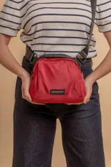 Bandolera pequeña de tela tafeta color rojo, con correa ancha ajustable de tejido negro y ribetes negros. Presenta un parche rectangular con el logo "LINCOLN'S" en el frente.