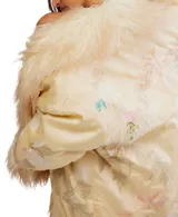 Chaqueta estilo kimono o bata, color crema o amarillo pálido, con acabado satinado y bordados florales sutiles. Presenta un ribete y puños voluminosos de pelo sintético largo y rizado en tono beige/crema.