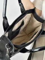 Bolso negro de cuero con detalles charolados, doble asa corta y correa larga de tela con el nombre de la marca.