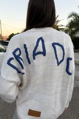 Buzo de tejido de punto color crudo, con cuello redondo y manga larga. Presenta un bordado frontal grande con un diseño abstracto que simula un rostro con rayos solares.