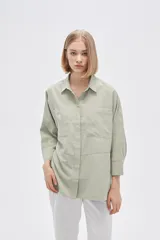 Camisa color verde claro, de corte holgado, con cuello clásico, cierre frontal con botones, mangas 3/4 y dos bolsillos de parche en el pecho.