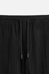 Pantalón negro de corte amplio, con textura acanalada, cintura elástica ajustable con cordón, bolsillos laterales y traseros de vivo.