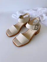 Sandalias de cuero color crudo, con tira ancha sobre los dedos, pulsera al tobillo con hebilla metálica y taco de madera de 6 cm de altura.