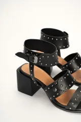 Sandalia de cuero vacuno color negro con tachas plateadas, tira ancha sobre los dedos, pulsera al tobillo con hebilla y taco cuadrado.