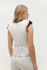 Chaleco blanco con finas rayas verticales negras, escote en V, cierre frontal con botones negros y volados en lino negro en los hombros. La parte inferior presenta otro volado en la misma tela del cuerpo.