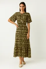 Vestido midi de manga corta, con estampado ikat en tonos verde militar y beige. Presenta cuello con abertura en V anudada con borlas colgantes y elástico fruncido en la cintura. La falda es escalonada.