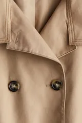 Gabardina corta color beige con doble botonadura, cuello clásico, charreteras en los hombros, bolsillos delanteros con solapa, mangas anchas con trabillas ajustables y pliegue en la espalda.