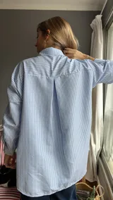 Camisa celeste a rayas verticales blancas, de corte holgado, con cuello clásico, mangas largas con ajuste de lazos y detalle de volados en los hombros.