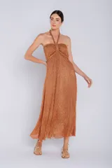 Vestido largo con escote halter anudado al cuello, confeccionado en tejido metalizado plisado color cobre/naranja con brillo. El corpiño presenta un fruncido central y la falda tiene una caída fluida.