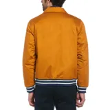 Campera estilo universitario color mostaza, con cierre de botones a presión, bolsillos laterales y un bolsillo interno. Tiene un parche con la letra P en el pecho y ribetes a rayas en puños y cintura.