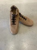 Zapatilla deportiva tipo 'champion' de gamuza color beige, con cordones marrones y cuello fruncido elástico. Presenta un diseño minimalista y liviano, con suela plana y taco escondido.