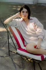 Buzo color crema con cuello polo, mangas largas y puños ajustados. Presenta un bolsillo en el pecho con bordados de frutas y la inscripción "Bom Faro by Coppelia". Los bordes del cuello, el bolsillo y los puños están decorados con puntadas rojas.