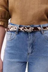 Cinturón de cuero con estampado animal print de vaca en tonos marrón y blanco, con hebilla metálica plateada.