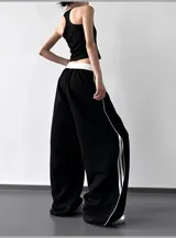 Pantalón deportivo negro de calce extra amplio (wide leg) con cintura elástica blanca y cordón ajustable blanco. Presenta tres tiras verticales blancas a lo largo de las costuras laterales.
