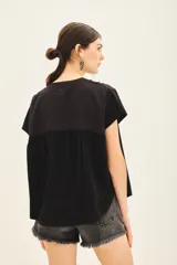 Camisa negra de manga corta con combinación de tejido de punto y plano, detalles de volados y frunces en pechera y espalda, acceso con botones fake nacar y medallita marcaria en espalda.