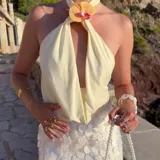 Top color crema o amarillo pálido, estilo halter con cuello atado y escote pronunciado en V con drapeado frontal. El cuello está adornado con una flor de orquídea en tonos amarillo, naranja y rosa.