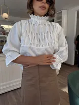 Blusa blanca de lino con cuello alto fruncido, mangas 3/4 con volados y detalle constructivo en el delantero con costuras a mano.