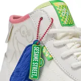 Zapatillas de caña alta Vans x Sesame Street, color blanco con detalles en rosa, verde y celeste. Presentan un estampado del personaje de la Rana René en el lateral y un dije de felpa con la letra L.