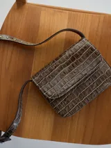 Cartera bandolera marrón con textura tipo piel de cocodrilo, solapa con cierre imantado y correa fina regulable.