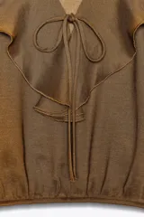 Blusa color marrón con escote en pico, manga larga abullonada con elástico en los puños y volantes en el escote y mangas.