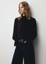 Blusa negra de tejido fluido con cuello alto, cierre frontal oculto con botones, mangas largas con puños elásticos y volantes.