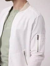 Campera bomber color blanco confeccionada en lino y algodón, con cuello mao, cierre frontal con cremallera, bolsillos frontales con cierre y puños y ruedo acanalados en color beige.