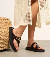 Sandalias planas de PU color marrón oscuro, con tira entre los dedos y tira ancha sobre el empeine con hebilla dorada ajustable.