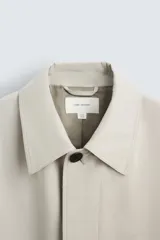 Gabardina color arena de corte recto, con cuello solapa, cierre frontal con botones, mangas largas con puños abotonados, bolsillos laterales y abertura en la parte posterior.