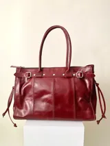 Bolso grande de cuero marrón con textura tipo cocodrilo, estructura rígida y asas para llevar al hombro. Tiene una tira con tachas y hebillas en cada extremo, también lleva lazos en los costados.