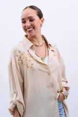 Camisa color beige con bordado de lentejuelas doradas en los hombros, corte clásico, cuello camisero y mangas largas con puños abotonados.