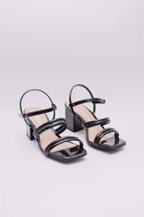 Sandalias de ecocuero charolado color negro, con punta cuadrada, dos tiras horizontales sobre el empeine, pulsera con hebilla al tobillo y taco cuadrado.