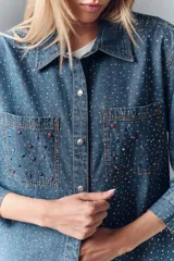 Camisa de denim azul con apliques metálicos plateados, de calce amplio, cuello camisero, cierre frontal con botones a presión, mangas largas con puños abotonados y dos bolsillos con solapa abotonada en el pecho.