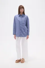 Camisa de algodón blanca de corte holgado, con cuello camisa y manga larga. Presenta un estampado de finas rayas verticales negras y un bolsillo frontal con rayas horizontales.