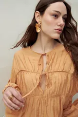 Blusa color naranja de caída fluida con cuello redondo y lazos frontales. Diseño con cortes horizontales y frunces, mangas largas con puños elastizados.