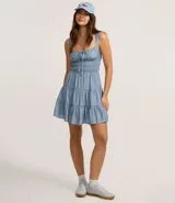 Vestido corto color beige, con escote recto, breteles medianos y lazo decorativo en el centro del busto. Presenta costura fruncida en la cintura y falda con babados en capas.