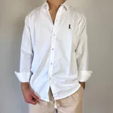 Camisa blanca de algodón con cuello clásico, manga larga con puños abotonados y cierre frontal con botones. Tiene un pequeño logo bordado en el pecho.