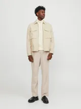 Chaqueta color crema, de manga larga, con cuello camisero y cierre frontal con cremallera metálica. Tiene dos bolsillos de parche con solapa en el pecho.