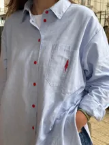 Camisa celeste de tela oxford, corte oversized, con bolsillo en el frente con bordado de árbol rojo y botones rojos.