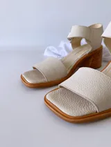 Sandalias de cuero color crudo, con tira ancha sobre los dedos, pulsera al tobillo con hebilla metálica y taco de madera de 6 cm de altura.