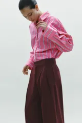 Camisa oversize rosada con rayas verticales blancas y celestes, cuello solapa, manga larga con puños abotonados, bolsillo de plastrón en el pecho y pinza en la espalda.