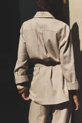 Camisa color marrón con cuello solapa y manga larga acabada en vuelta con trabilla y botón. Bolsillos delanteros de plastrón con solapa. Bajo con aberturas laterales. Cierre frontal con botones y cinturón de la misma tela.