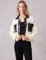 Chaqueta de corte boxy en color crema, confeccionada en tejido de punto suave con detalles en cuero sintético negro en cuello, solapas, cartera frontal y bolsillos. Presenta cierre frontal con botones metálicos plateados, hombros caídos y costuras decorativas horizontales.